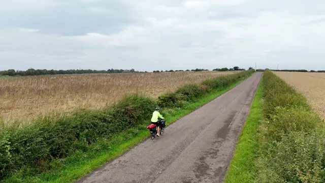 Amy riding a loaded Brompton down a narrow lane between hedges and a barley field