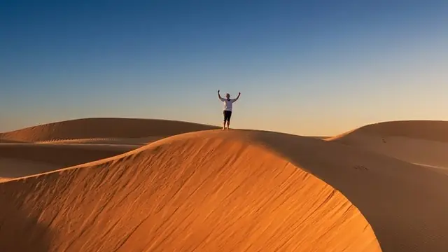 A small figure with arms raised on the crest of an orange sand dune, blue sky above
