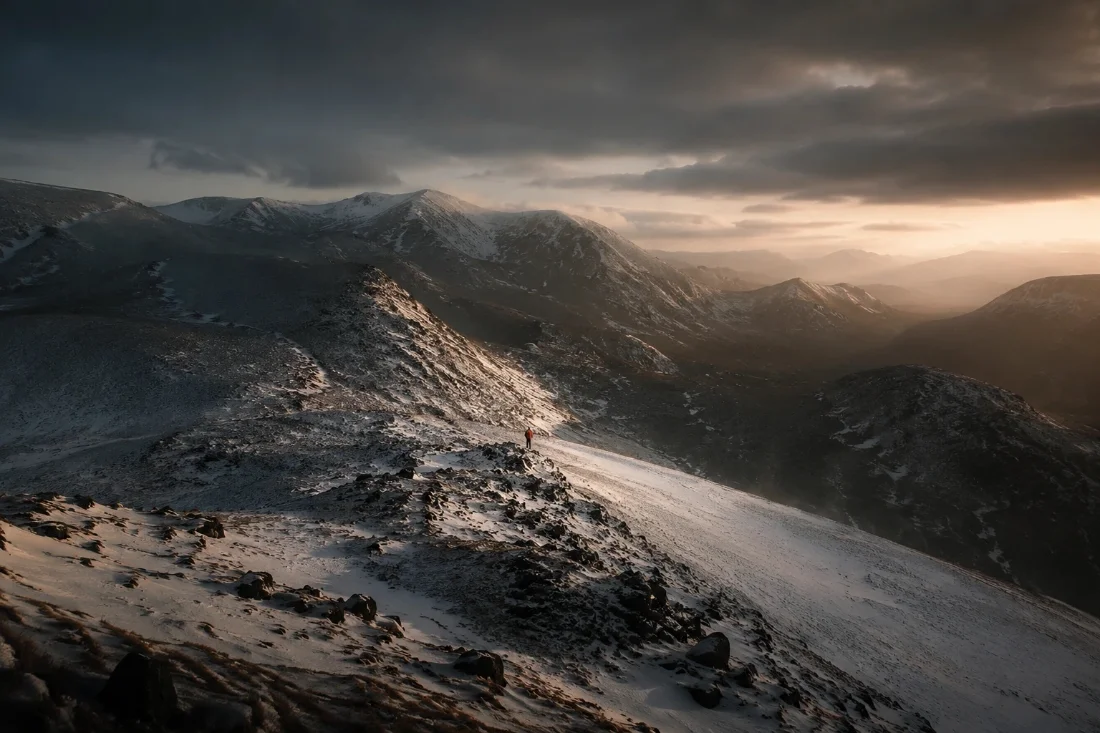 A lone walker in a red jacket on a snowy ridge as storm light breaks gold across the hills